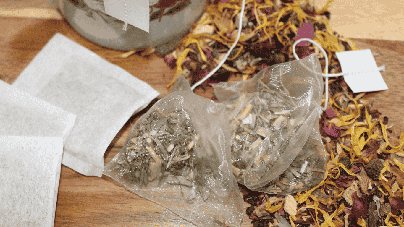 A range of tea bags some containing plastics and some not, along with some loose leaf tea sits on a wood background next to a coffee cup.