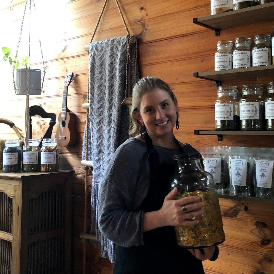 A woman standing in a herbal apothecary holding a jar of yellow Calendula flowers.