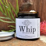 Whip Pure Tallow Balm Australia sits on a stone base with timber background. Decorative Australian native flowers in the background.