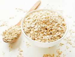 A white bowl overflowing with oats. A wooden spoon sits beside it on a white background.