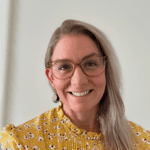 Asti Mary Sommerfield, founder of My Herbal Apothecary, smiling warmly. She is wearing glasses and a yellow floral blouse, with light blonde hair styled to one side. The background is a simple, neutral-colored wall.