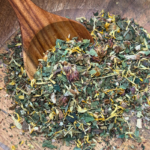 A beautifully crafted mango wood bowl filled with Radiance Organic Herbal Tea sits on a natural surface. The loose leaf blend, featuring vibrant calendula petals, rooibos, and nourishing herbs, is visible in rich detail. A wooden spoon rests beside the bowl, ready for scooping, complementing the earthy, handcrafted aesthetic of the scene.