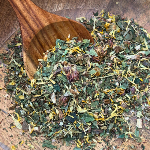 A beautifully crafted mango wood bowl filled with Radiance Organic Herbal Tea sits on a natural surface. The loose leaf blend, featuring vibrant calendula petals, rooibos, and nourishing herbs, is visible in rich detail. A wooden spoon rests beside the bowl, ready for scooping, complementing the earthy, handcrafted aesthetic of the scene.