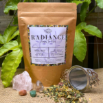 A kraft paper bag of Radiance Organic Herbal Tea sits on a natural surface, surrounded by loose leaf tea showcasing vibrant calendula petals, rooibos, and herbs. A stainless steel tea infuser rests nearby, ready for steeping. Polished crystals add a touch of serenity, with a leafy passionfruit leaf in the background, enhancing the natural and nourishing essence of the blend.