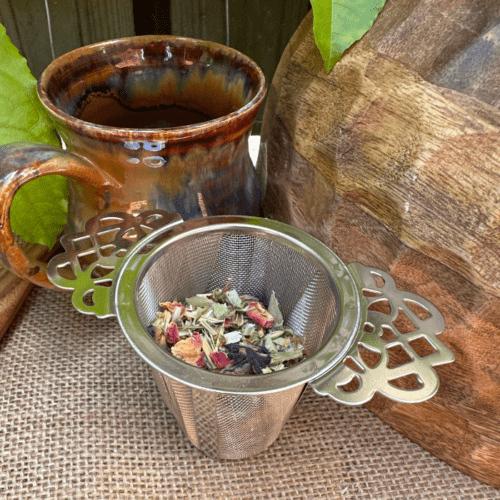 A Serenity Herbal Tea Infuser (loose leaf tea strainer) rests in a handmade ceramic mug, set on a hessian background with fresh passionfruit leaves. Beside the mug, a mango wood bowl and a handcrafted wooden spoon create an inviting scene. The infuser is filled with loose leaf Sorrow Organic Herbal Tea, showcasing a vibrant blend of herbs, petals, and botanicals. The setting evokes a sense of natural tranquility and mindfulness.