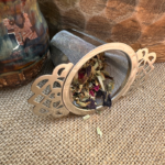A Serenity Herbal Tea Infuser (loose leaf tea strainer) rests next to a handmade ceramic mug, set on a hessian background. Beside the mug, a mango wood bowl creates an inviting scene. The infuser is filled with loose leaf Sorrow Organic Herbal Tea, showcasing a vibrant blend of herbs, petals, and botanicals. The setting evokes a sense of natural tranquility and mindfulness.