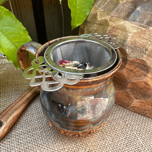 A Serenity Herbal Tea Infuser (loose leaf tea strainer) rests in a handmade ceramic mug, set on a hessian background with fresh passionfruit leaves. Beside the mug, a mango wood bowl and a handcrafted wooden spoon create an inviting scene. The infuser is filled with loose leaf Sorrow Organic Herbal Tea, showcasing a vibrant blend of herbs, petals, and botanicals. The setting evokes a sense of natural tranquility and mindfulness.