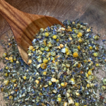 A beautifully crafted mango wood bowl filled with Slumber Organic Herbal Tea sits on a neutral-toned surface. The loose leaf blend of chamomile, lavender, spearmint, and fennel seeds is visible, showcasing its delicate textures and earthy colors. A wooden spoon rests beside the bowl, ready for scooping, adding to the warm, natural aesthetic of the scene.
