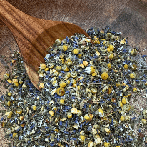 A beautifully crafted mango wood bowl filled with Slumber Organic Herbal Tea sits on a neutral-toned surface. The loose leaf blend of chamomile, lavender, spearmint, and fennel seeds is visible, showcasing its delicate textures and earthy colors. A wooden spoon rests beside the bowl, ready for scooping, adding to the warm, natural aesthetic of the scene.