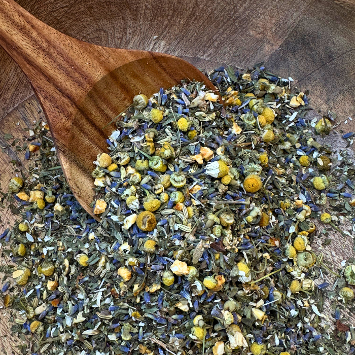 A beautifully crafted mango wood bowl filled with Slumber Organic Herbal Tea sits on a neutral-toned surface. The loose leaf blend of chamomile, lavender, spearmint, and fennel seeds is visible, showcasing its delicate textures and earthy colors. A wooden spoon rests beside the bowl, ready for scooping, adding to the warm, natural aesthetic of the scene.