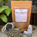 A kraft paper bag of Slumber Organic Herbal Tea sits on a natural linen surface, with loose leaf tea scattered beside it. The blend of chamomile, lavender, spearmint, and fennel seeds is visible among the dried herbs. A stainless steel tea strainer rests nearby, ready for steeping. Small, polished crystals add a touch of tranquility, enhancing the calming aesthetic of the scene.