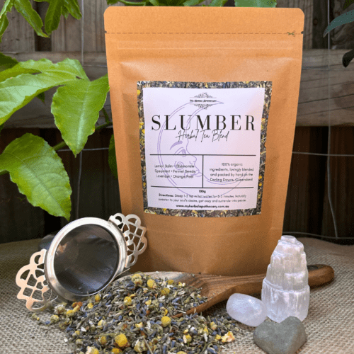 A kraft paper bag of Slumber Organic Herbal Tea sits on a natural linen surface, with loose leaf tea scattered beside it. The blend of chamomile, lavender, spearmint, and fennel seeds is visible among the dried herbs. A stainless steel tea strainer rests nearby, ready for steeping. Small, polished crystals add a touch of tranquility, enhancing the calming aesthetic of the scene.