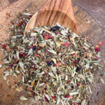 A kraft paper bag of Sorrow Organic Herbal Tea rests on a hessian base with leafy passionfruit leaves in the background. Loose leaf herbal tea, showcasing a blend of hawthorn, oat straw, rose petals, and butterfly pea flower, spills gently over the edge of a wooden spoon. A stainless steel tea infuser is nearby, ready for steeping, evoking a sense of heart-centered comfort and reflection.