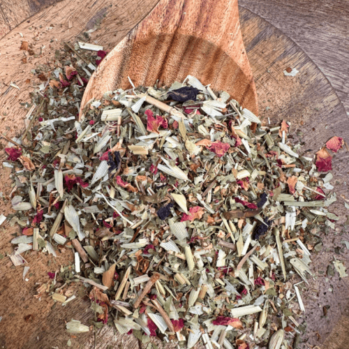 A kraft paper bag of Sorrow Organic Herbal Tea rests on a hessian base with leafy passionfruit leaves in the background. Loose leaf herbal tea, showcasing a blend of hawthorn, oat straw, rose petals, and butterfly pea flower, spills gently over the edge of a wooden spoon. A stainless steel tea infuser is nearby, ready for steeping, evoking a sense of heart-centered comfort and reflection.