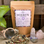 A kraft paper bag of Sorrow Organic Herbal Tea rests on a hessian base with leafy passionfruit leaves in the background. Loose leaf herbal tea, showcasing a blend of hawthorn, oat straw, rose petals, and butterfly pea flower, spills gently over the edge of a wooden spoon. A stainless steel tea infuser is nearby, ready for steeping, evoking a sense of heart-centered comfort and reflection.