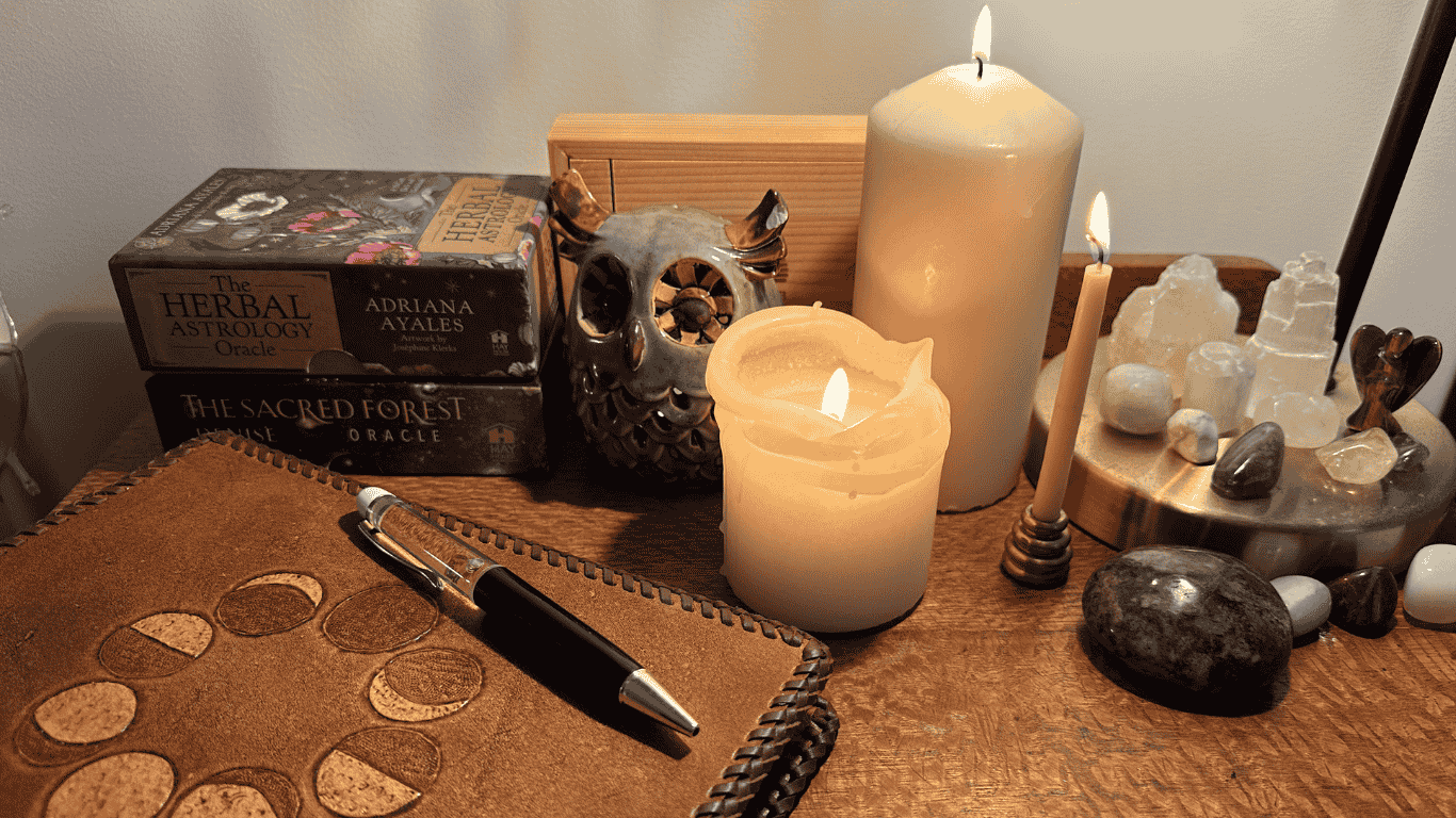 A calming ritual setup with a leather moon phase journal and pen, oracle decks, lit candles, a ceramic owl, and a crystal display on a wooden surface — creating a warm, reflective space for healing and self-care.