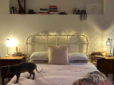 A warm, softly lit bedroom with fairy lights above an iron bed frame, flanked by wooden bedside tables holding books, candles, and crystals. A hairless cat stands on the quilted weighted blanket, adding a sense of comfort and presence.