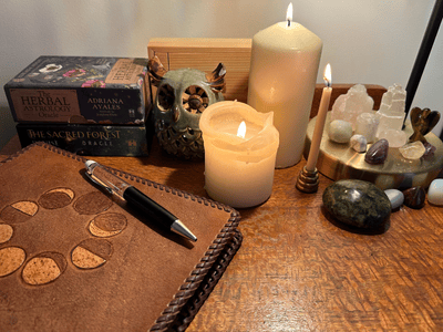 A cozy ritual space with lit candles, crystal clusters, an owl-shaped incense holder, and stacked oracle card decks. A leather-bound journal with a moon phase design and a pen rest on a wooden surface, creating a calming setting for reflection or spiritual practice.