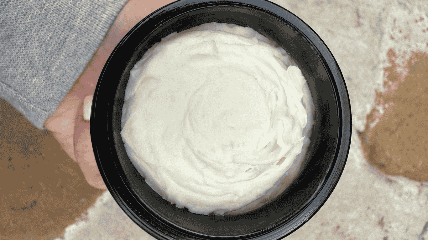 A close-up of a jar filled with creamy whipped beef tallow balm for skin, held in a hand against a textured natural background.