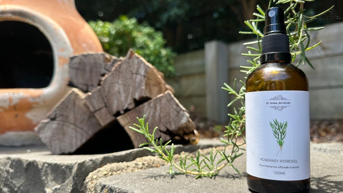 Bottle of My Herbal Apothecary Rosemary Hydrosol sitting outdoors among fresh rosemary sprigs, with firewood and a garden chiminea in the background.